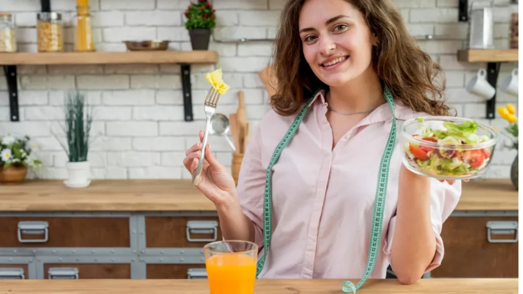 Exemplo de Desintoxicação Mulher segurando uma tijela de vidro com verduras cortadas dentro com a mão esquerda e um garfo com uma verdura espetada na mão direita