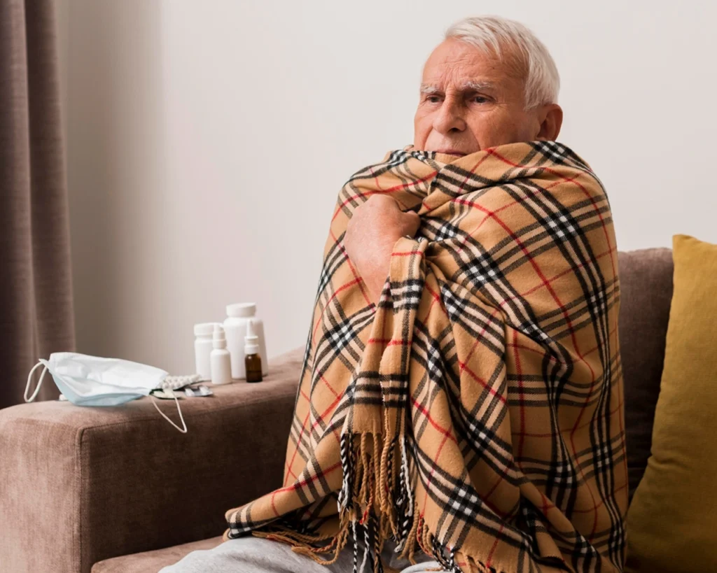 Homem sénior com gripe enrolado num cobertor, ao lado de máscara e medicamentos — representação realista da gripe depois dos 50 anos.
