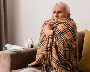 Homem sénior com gripe enrolado num cobertor, ao lado de máscara e medicamentos — representação realista da gripe depois dos 50 anos.
