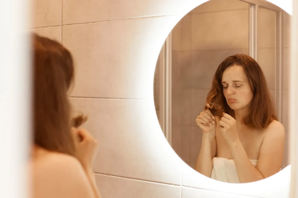 Mulher adulta preocupada com a queda de cabelo ao olhar para o espelho, segurando fios nas mãos, representando eflúvio telógeno e perda capilar.